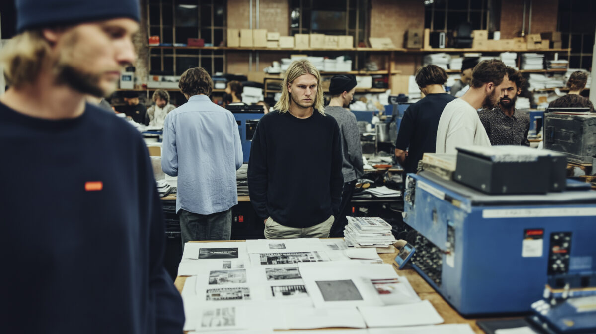 Multiple team members interact around workstations and printouts, depicting dynamic coordination in an office setting.