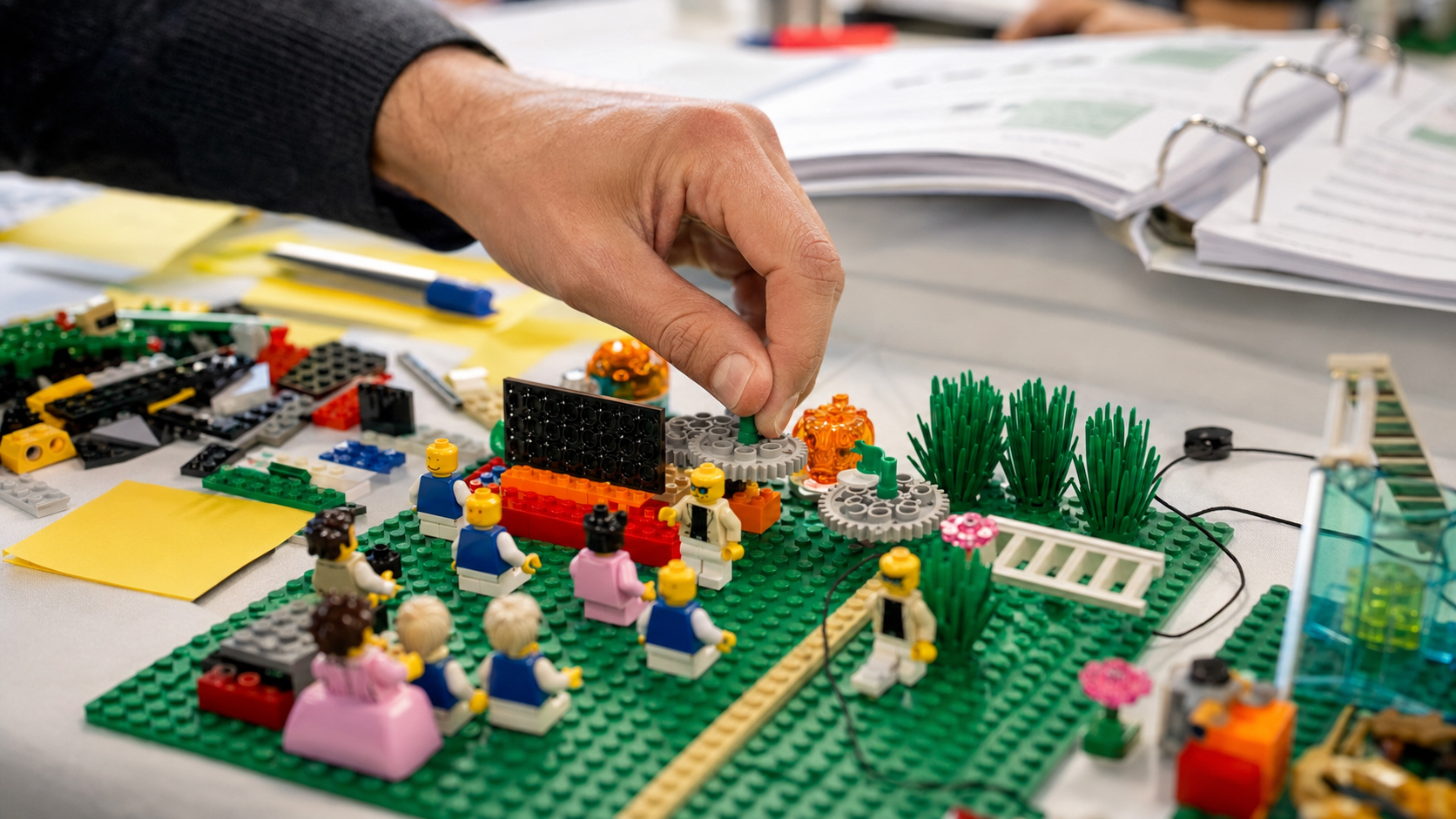 Facilitator’s hand adjusts a LEGO Serious Play workshop model with minifigures, gears, and bricks on a table.