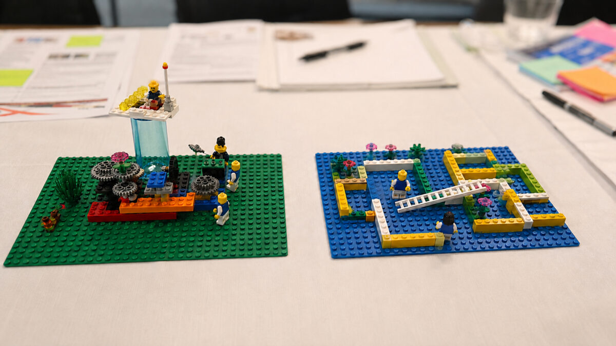 Two LEGO Serious Play workshop models on a table show different team perspectives during a facilitation session.