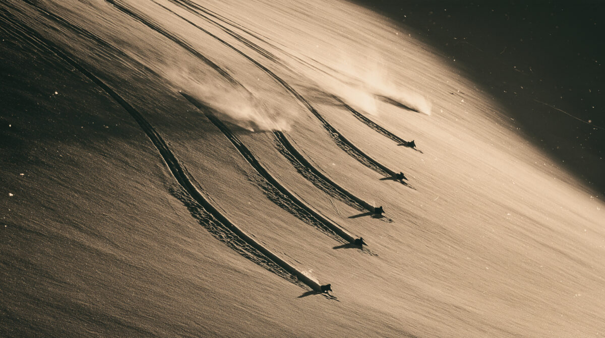 Aerial view of five skiers in silhouette descending a sunlit snow slope, each leaving a separate parallel track.