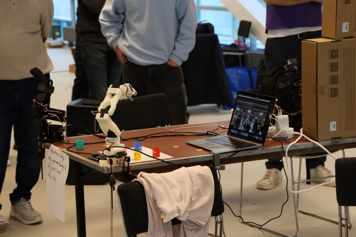 Hackathon demo table with a white robotic arm, coloured slinkies, a laptop and stacked robot kit boxes, people standing behind.